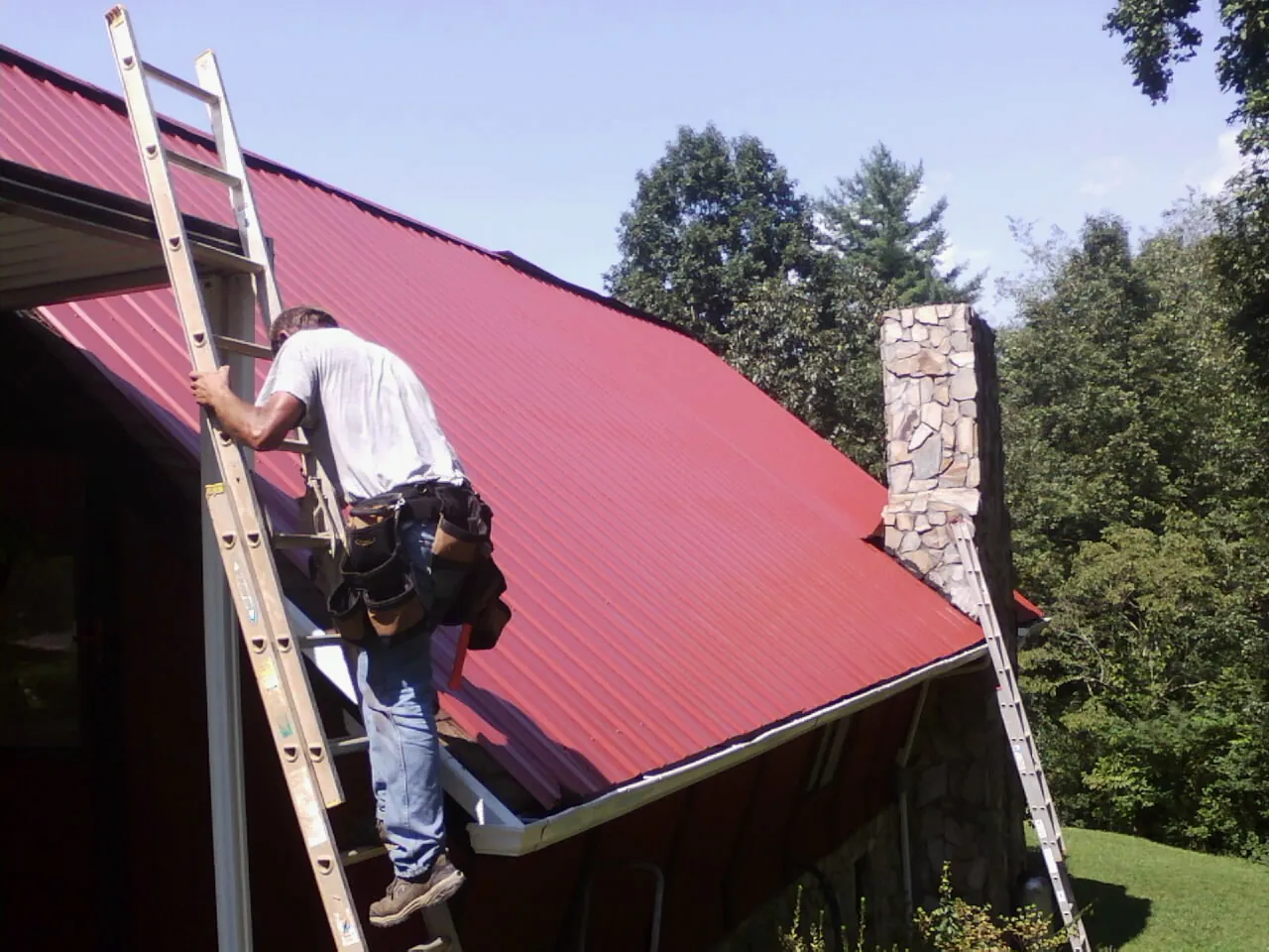 Roofer on a ladder beside a red metal roof and stone chimney — Soffit & Fascia Repair by Roofing Brothers in Sperryville
